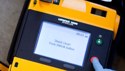 First responder pressing the red shock button on the LIFEPAK 1000 AED while the screens reads "Stand clear! Push the SHOCK button"